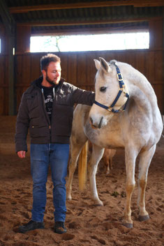 Ein stattlicher junger Mann mit Bart steht neben einen weißen Araber-Pferd und streichelt es am Hals. Das Pferd wendet dem Mann interessiert und ruhig den Kopf zu. 