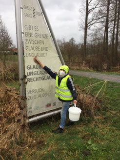 Eine Frau mit Warnweste und Eimer in der Hand reinigt ein Hinweisschild, das in Form eines Segels geformt ist.