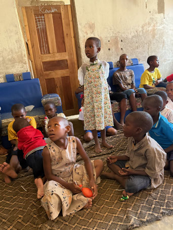 Mehrere Kinder sitzen oder stehen im Innenraum eines Hauses, teils mit Schüsseln auf dem Boden, offenbar vor der Essensausgabe