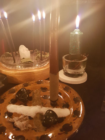 Verschiedene Kerzen die magisch angeordnet sind auf Tellern umgeben von Salz, Blüten, Kräuter und Steinen, die Kerzen leuchten hell und warm, sie befinden sich auf einem schwarzen altar