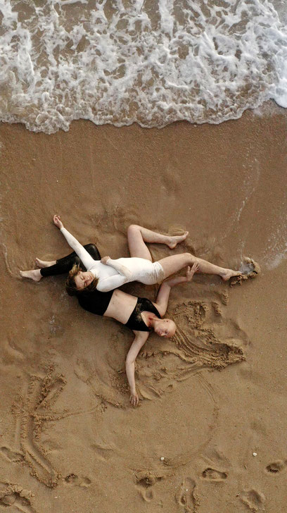 V3 Veruschka Bohn in weißem Body und Nina Blum in schwarz, sind von der Vogelperspektive fotografiert, während sie am Ufer eines Meeres ineinandergeknotet liegen, sich langsam entknoten, während die Wellen sie berühren..