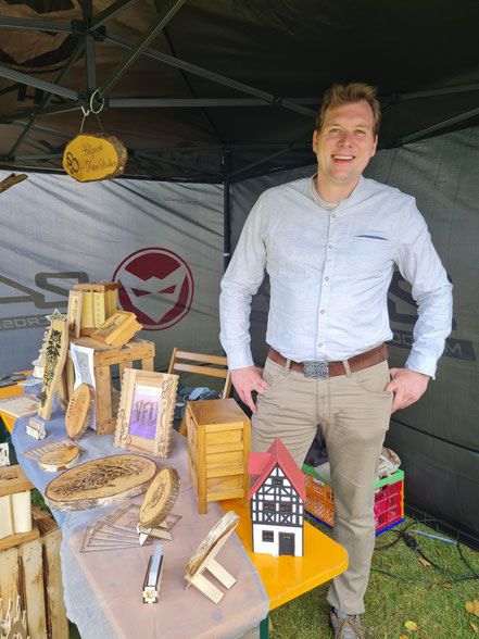 Ein lächelnder Mann steht an einem Messestand von Holzwerk Peter Stoiber unter einem Pavillon. Auf dem Tisch präsentiert er individuelle Holzkunst, kunstvolle Brandmalerei auf Baumscheiben, Holzboxen und ein Fachwerkhaus-Modell.