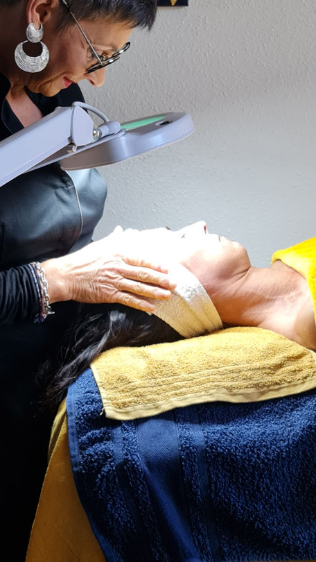 Une physio-esthéticienne effectue un diagnostic de peau sur une cliente allongée, à l’aide d’une lampe loupe. La cliente porte un bandeau en tissu sur le front et repose sur une table recouverte de serviettes jaunes et bleues. L’esthéticienne est concentr