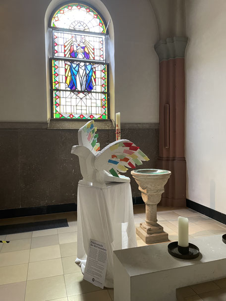 Eine Skulptur einer mit bunten Federn geschmückten, weißen Taube steht vor einem Glasfenster mit Marienmotiv. Dazwischen sind das Taufbecken und die Osterkerze zu erkennen.