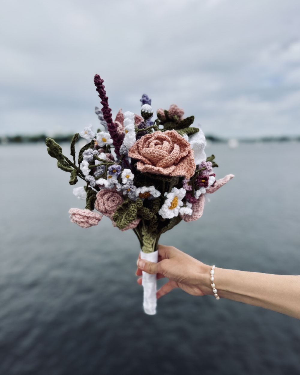 Ein selbst gehäkelter Blumenstrauss, Rosen, Kamille, Blätter, alles gehäkelt. Eine Hand hält ihn in die Höhe, im Hintergrund ist die Alster mit Segelbooten zu sehen