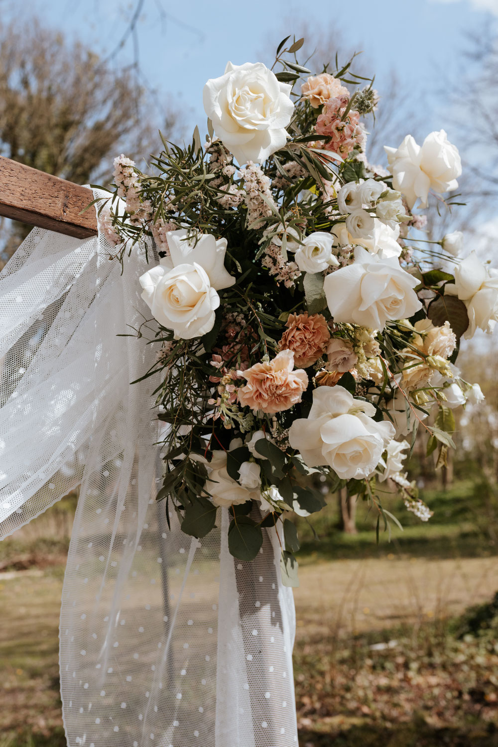 oppulente Blumendeko mit weißen Rosen, aprikotfarbenen Nelken und Eukalyptus am Traubogen. Der weiß gepunktete Stoff am Traubogen weht im Wind
