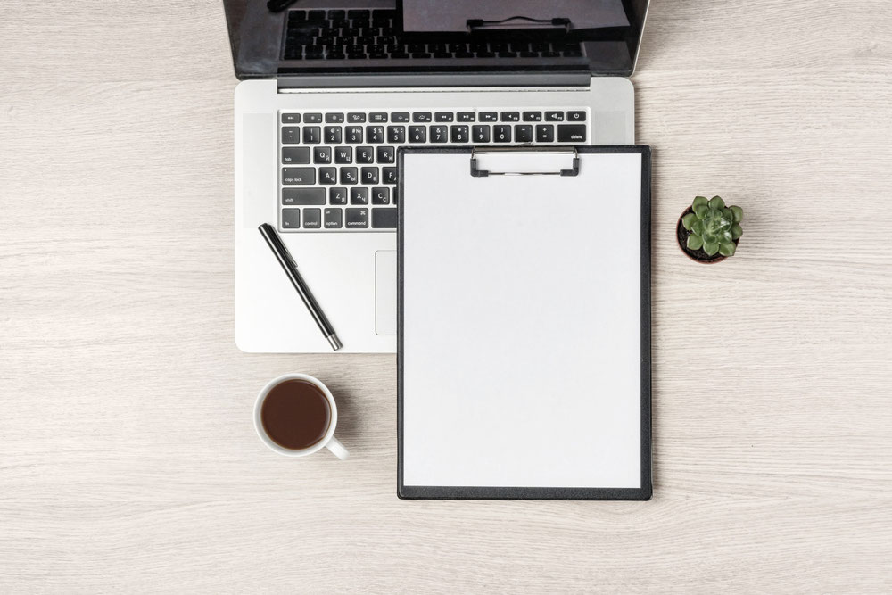 Ein Bild auf einem Schreibtisch mit Notebook, Stift, einem Klemmbrett, einer Tasse Kaffee und einer Pflanze