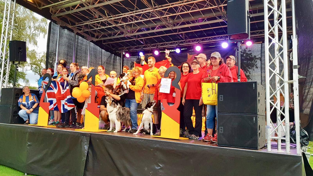 Mehrere Personen auf einer Bühne mit bunten T-Shirts, einige halten orangefarbene Luftballons, zwei Hunde sitzen vorne auf der Bühne.