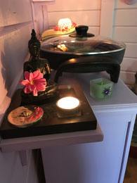 Idée de chaleur et de douceur : réchaud en fonte et son couvercle en verre ; et au 1er plan une petite bougie éclairant un bouddha assis ; ambiance pastelle chaude