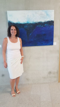 Miriam steht vor eines ihrer Acrylbilder in blau und weiß und lächelt in die Kamera
