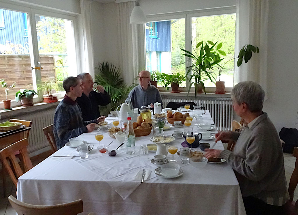Eine Gruppe von Menschen sitzt um eine Frühstückstafel in einem Tagungshaus und unterhält sich angeregt