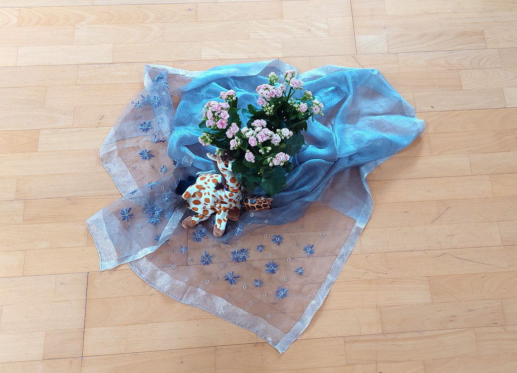Auf einem hellen Laminatfußboden ist ein bläulich schimmerndes, besticktes Tuch um einen kleinen Blumentopf drapiert. Daneben sitzt eine kleine Giraffe aus Plüsch.