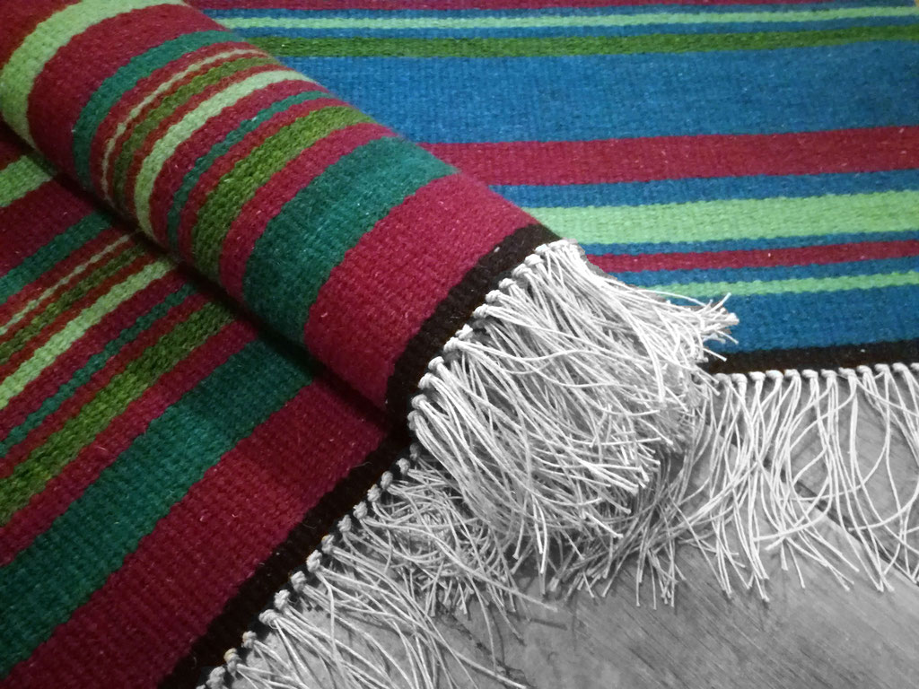 Deux tapis - descend de lit - rouge bordeaux et turquoise aux rayures irrégulières vertes sont posés sur le parquet en bois. Le tapis rouge est partiellement enroulé. Les franges en fils de lin son en premier plan 