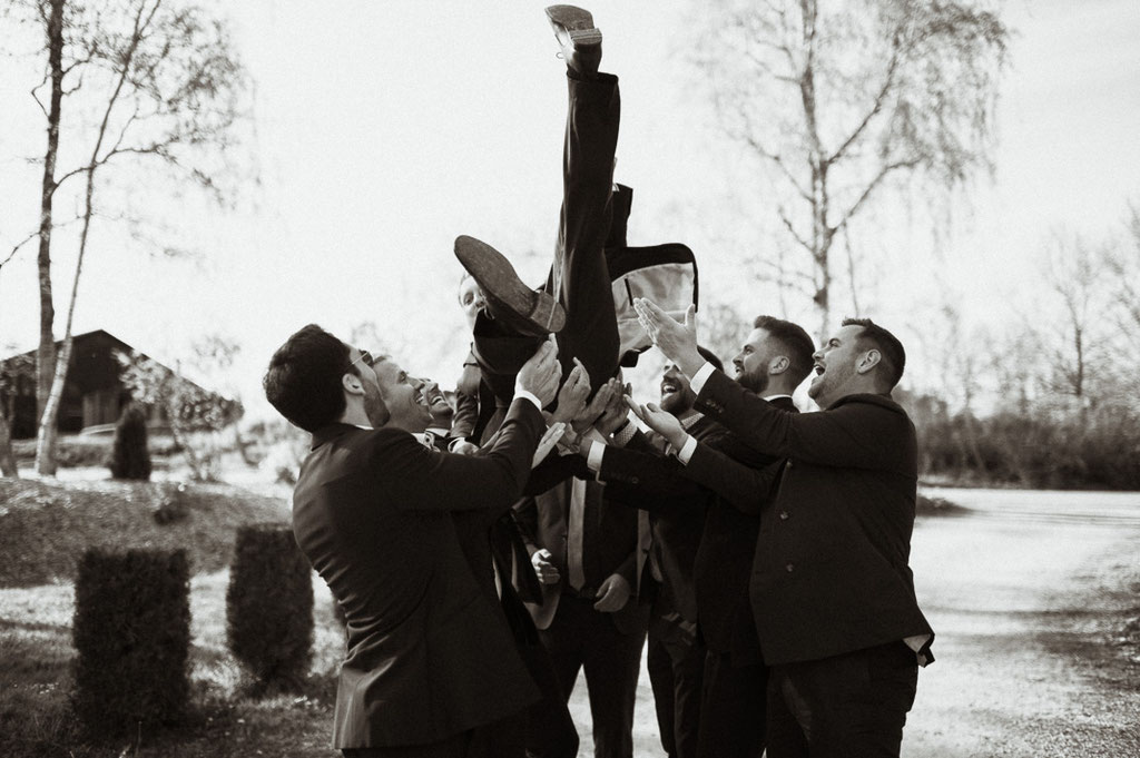 Der Bräutigam wird von seinen Freunden hochgeworfen. Ein spaßiges Hochzeitsfoto. Hochzeitsfotografen aus Freising.
