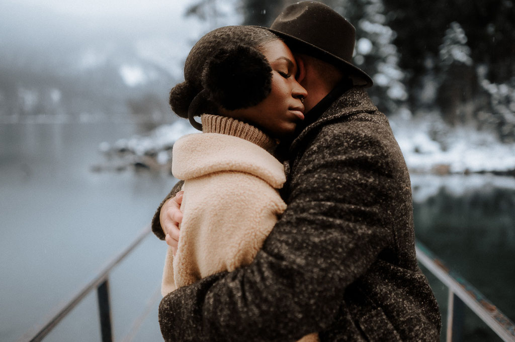 Ein Mann umarmt eine Frau beim Paarshooting, sie legt ihren Kopf auf seiner Schulter ab und hat die Augen geschlossen. Sie stehen auf einer Brücke beim winterlichen Eibsee.