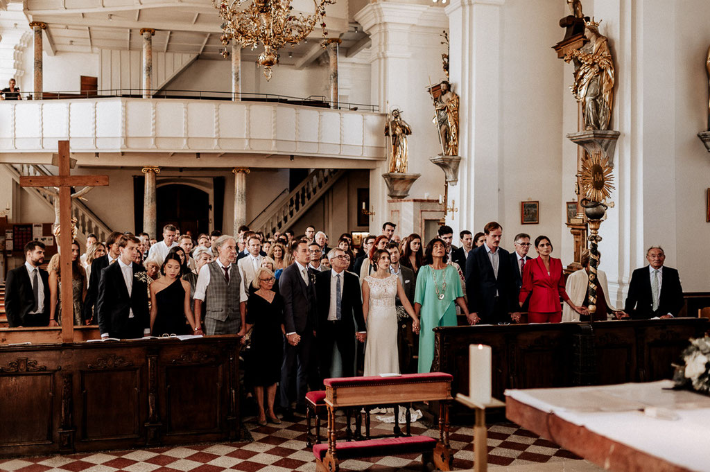 Hochzeitsfotograf in der St. Sixtus Kirche am Schliersee.Die Hochzeitsgesellschaft gibt sich zum Gebet die Hände. Reportagefoto von der Trauung.