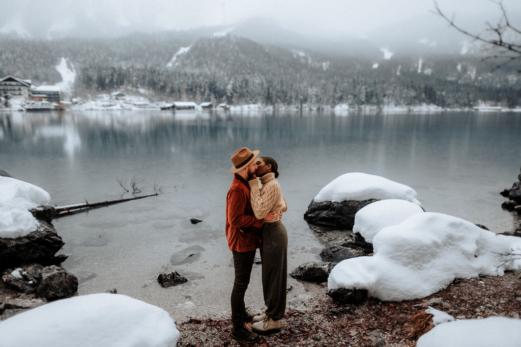 Ein Paar steht vor dem glasklaren Eibsee und küsst sich Im Hintergrund sind die Berge zu sehen.