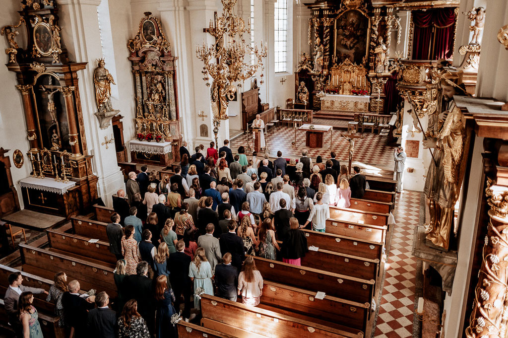 Hochzeitsfotograf in der St. Sixtus Kirche am Schliersee.Die Hochzeitsgesellschaft gibt sich zum Gebet die Hände. Reportagefoto von der Empore aus fotografiert.