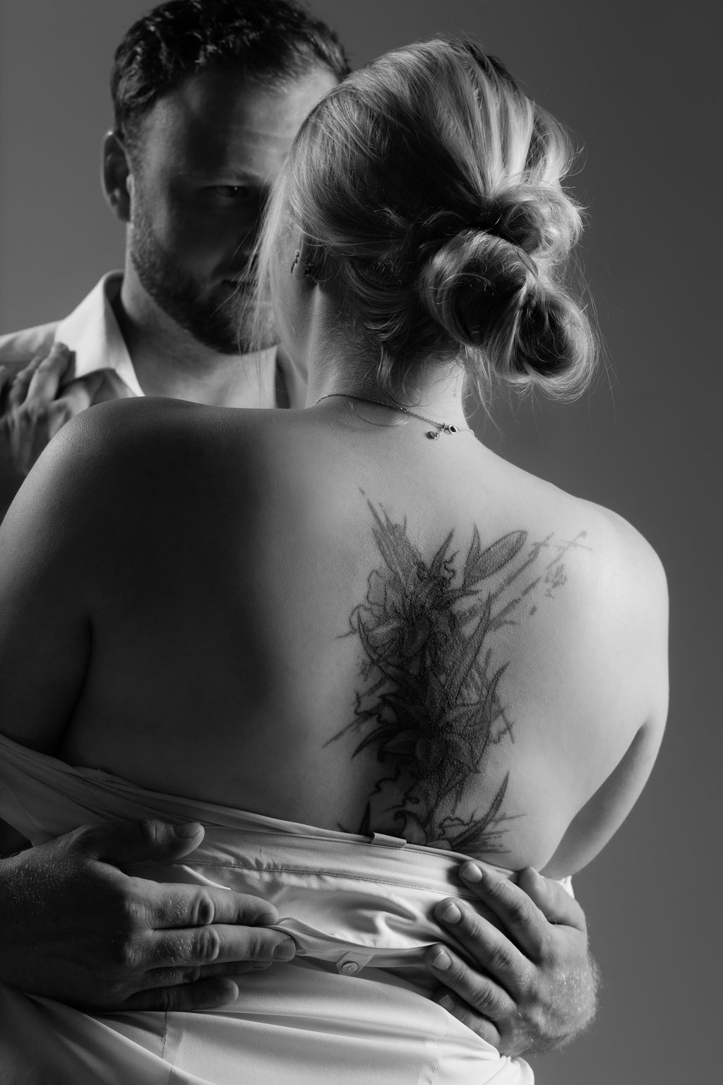 Sinnliche Paarfotografie im Studio: Umarmung, Frau mit Rückentattoo im Laken, Partner hält sie fest.