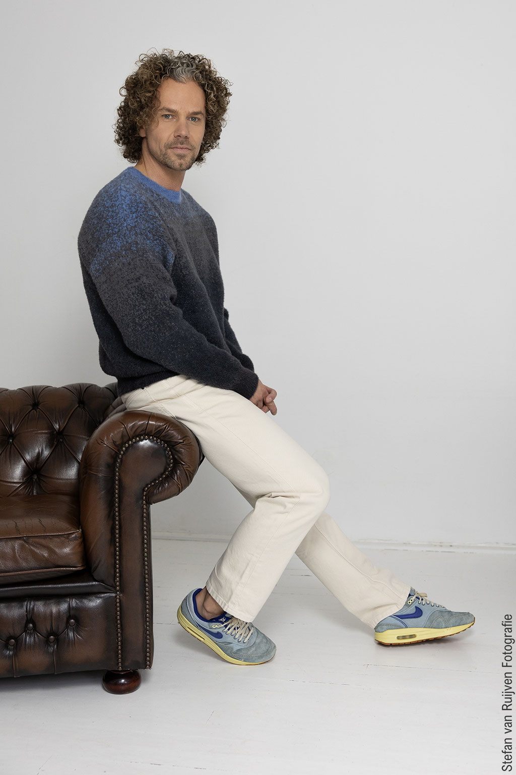 Stefan poseert op een bruine leren bank in Studio Duinlust. Portfolio maken tijdens de Fotoshoot Dag op vrijdag 27 februari 2026. Hij draagt een blauw met grijze trui, lichte broek en Nike sneakers in blauw/geel. Foto's gemaakt door Stefan van Ruijven