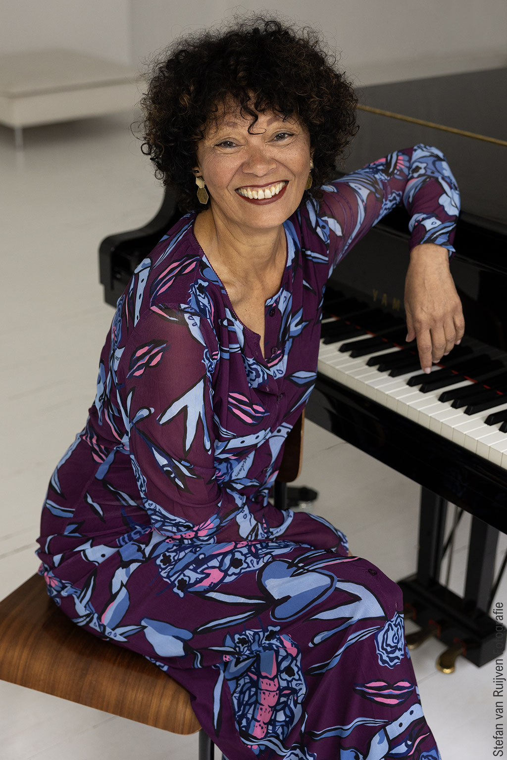 Yolanda zit in een paars met blauwe jurk met een  grote glimlach achter de piano op een stoel. De foto is gemaakt door Stefan van Ruijven tijdens de Fotoshoot Dag die hij als fotograaf heeft georganiseerd speciaal voor modellen en acteurs om hun portfolio