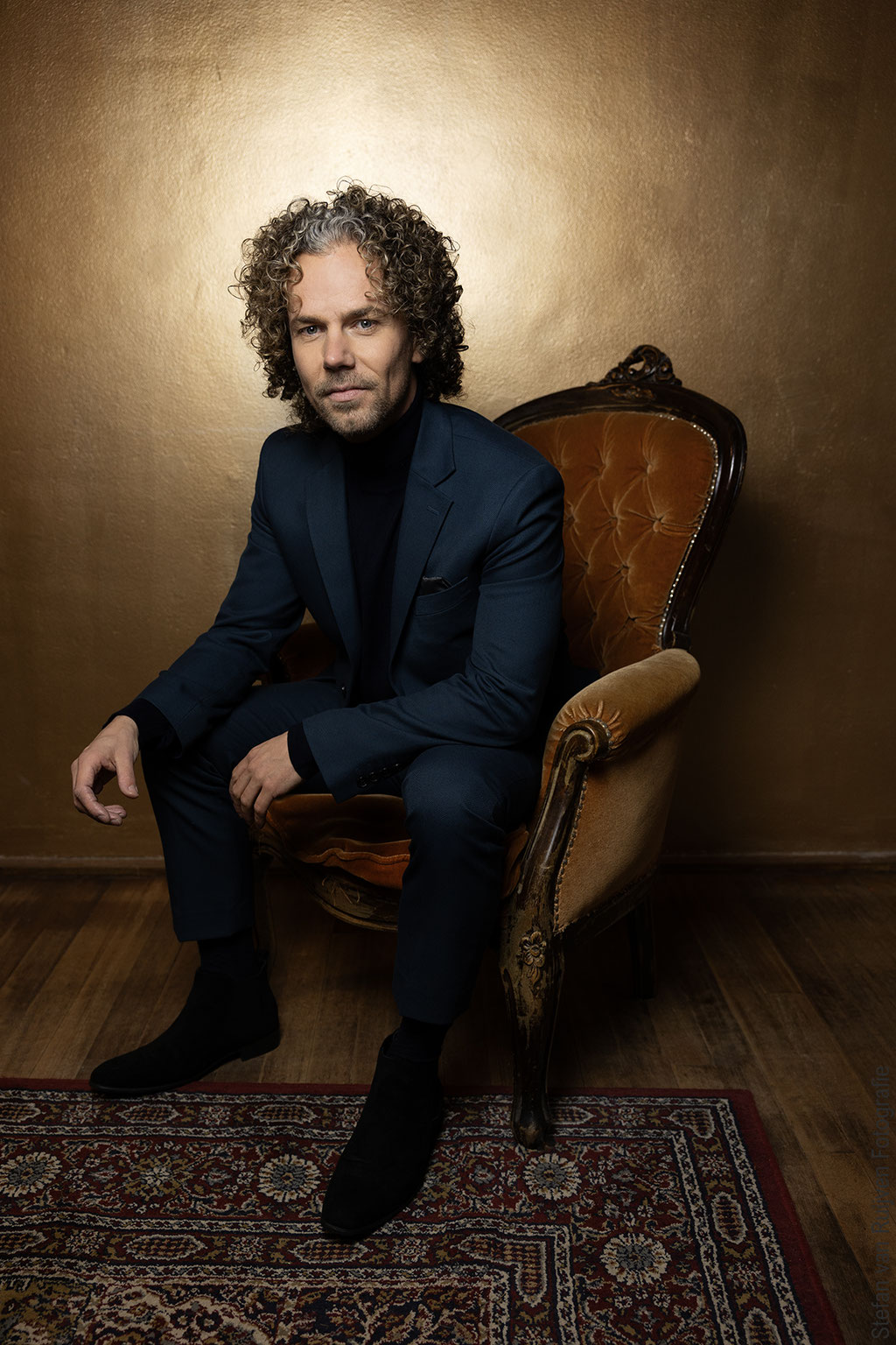 Stefan poseert in kamer met vintage kleed en stoel in Studio Duinlust. Portfolio maken tijdens de Fotoshoot Dag op vrijdag 27 februari 2026. Hij draagt een groen/blauw pak en zwarte coltrui. Foto's gemaakt door Stefan van Ruijven
