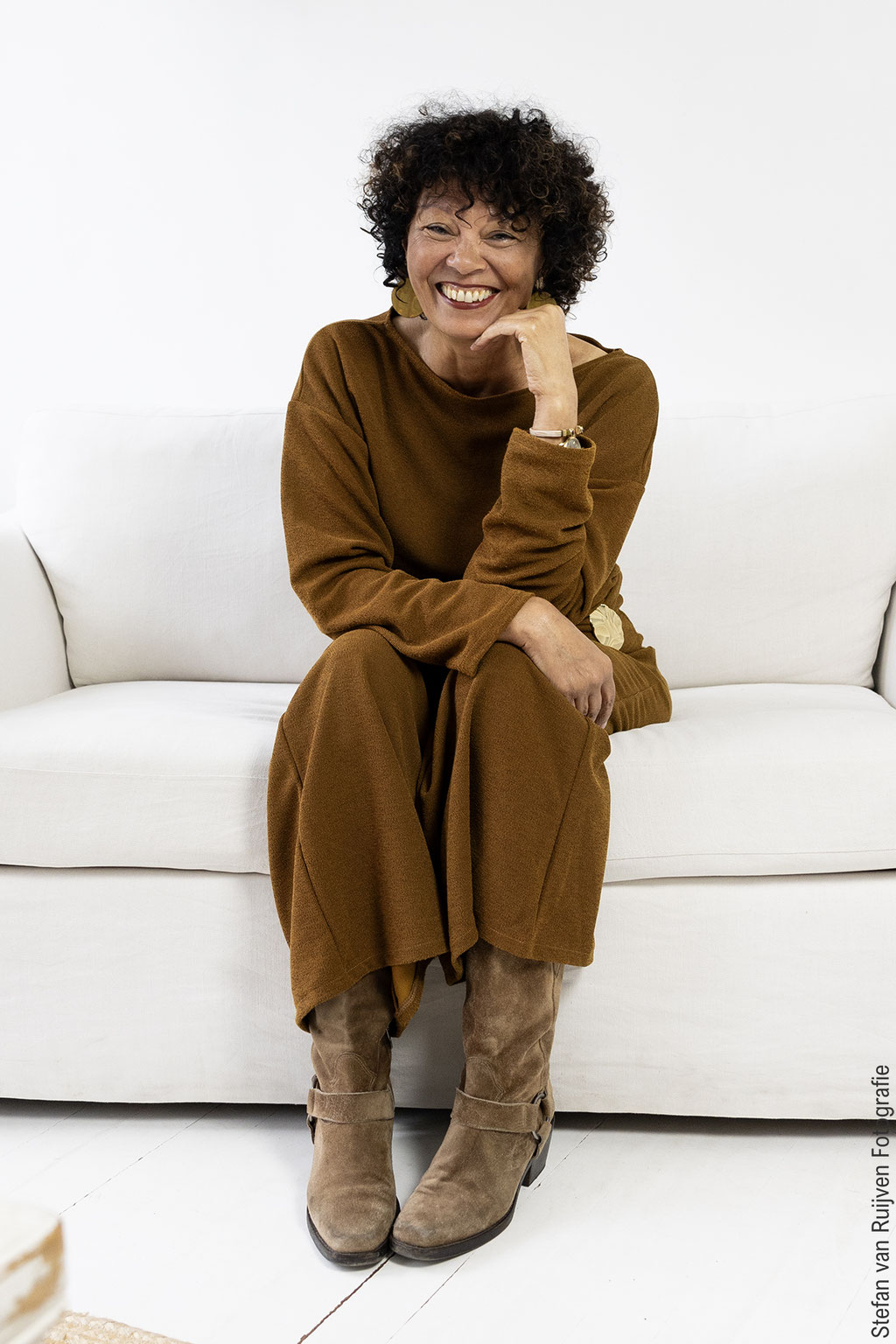 Yolanda zittend op een witte bank in Studio Duinlust. Ze draagt een bruine outfit met laarzen en geeft een grote glimlach. Deze foto is gemaakt tijdens de Fotoshot Dag in Studio Duinlust en is georganiseerd door Stefan van Ruijven Fotografie
