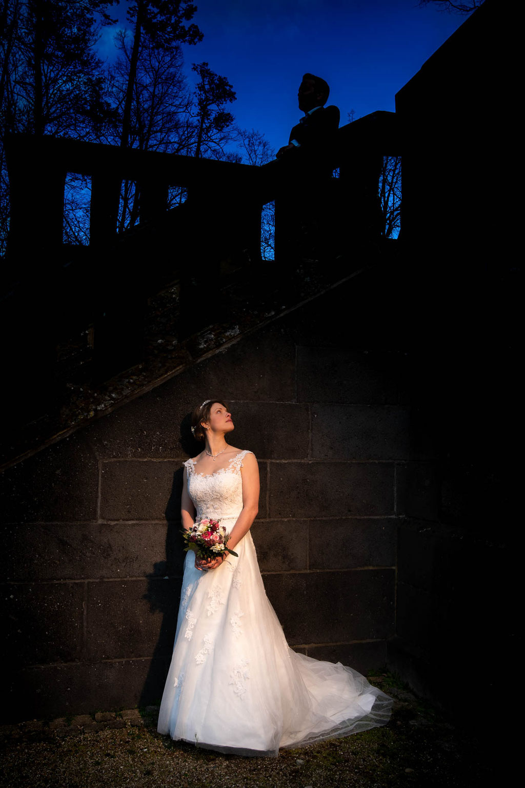 Hochzeitsfotograf Frankfurt - Braut steht im Scheinwerferlicht an Schloss Waldthausen in Budenheim an einer Treppe und Bräutigam steht in der Dunkelheit - special effect