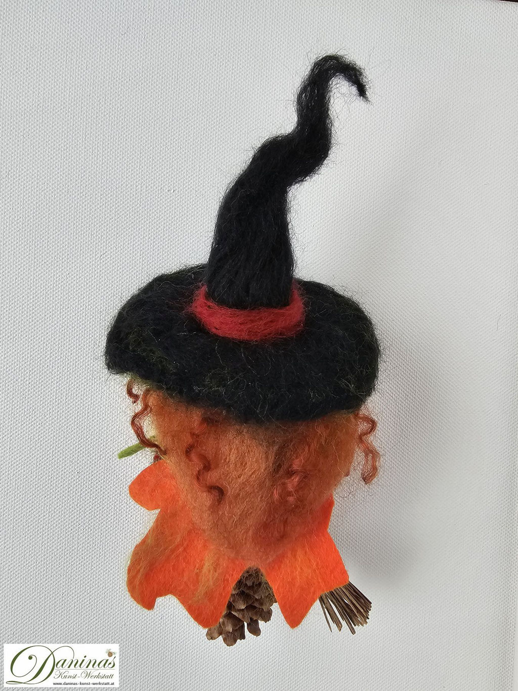 Hexe Amber Rückseite. Handgefertigte Märchenfigur aus Fichtenzapfen mit nadelgefilztem Kopf und rostroten langen Haaren aus Wolle, einem schwarz-grünen Hexenhut mit Zauberstein, einer Jacke aus Filz in Herbstfarben und einem Hexenbesen