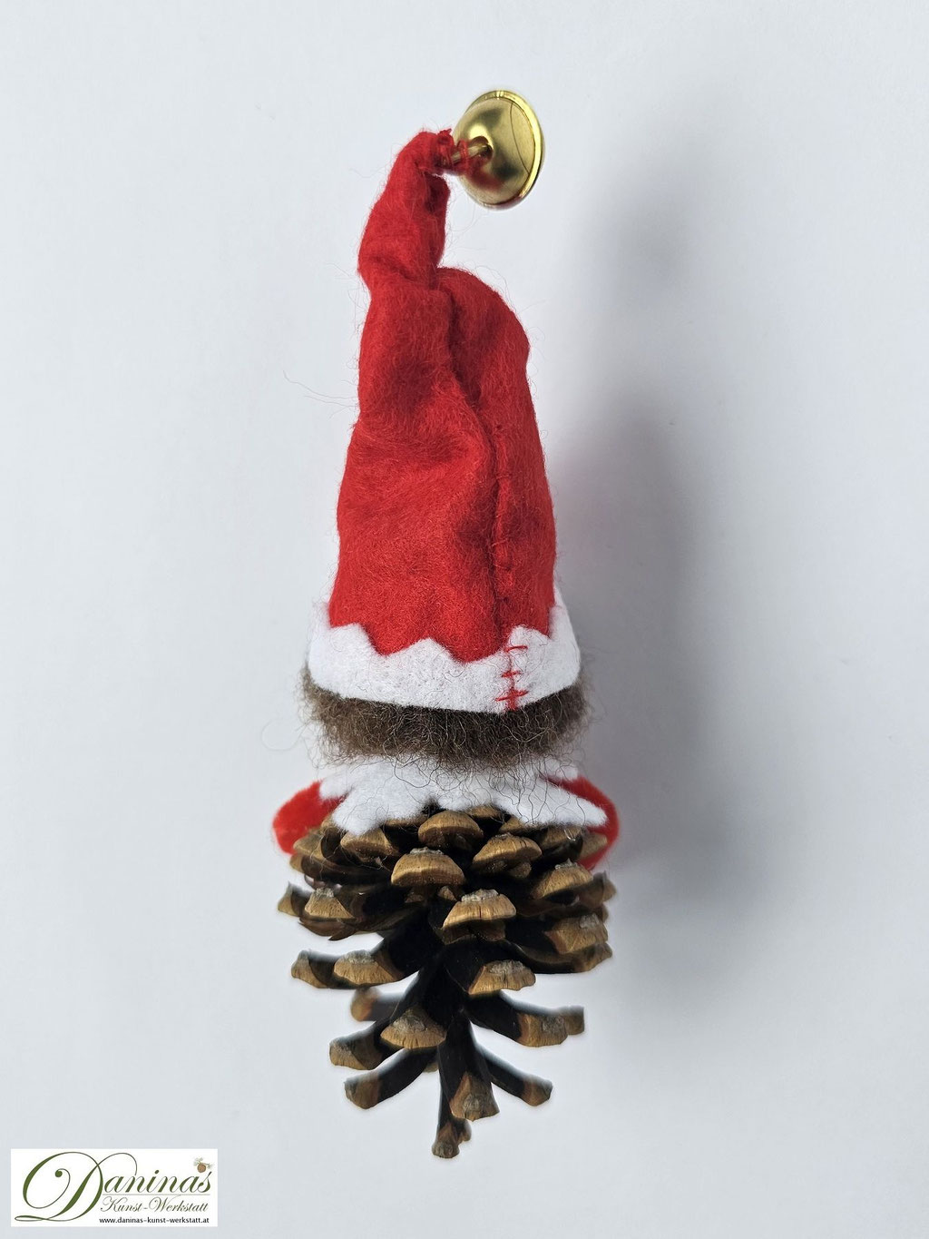 Weihnachtswichtel Floki, Rückseite. Handgemachte Wichtelfigur aus Kiefernzapfen mit braunen Haaren aus Wolle, rot-weißer Filz-Zipfelmütze mit goldfarbigem Glöckchen, weißem Filz Kragen