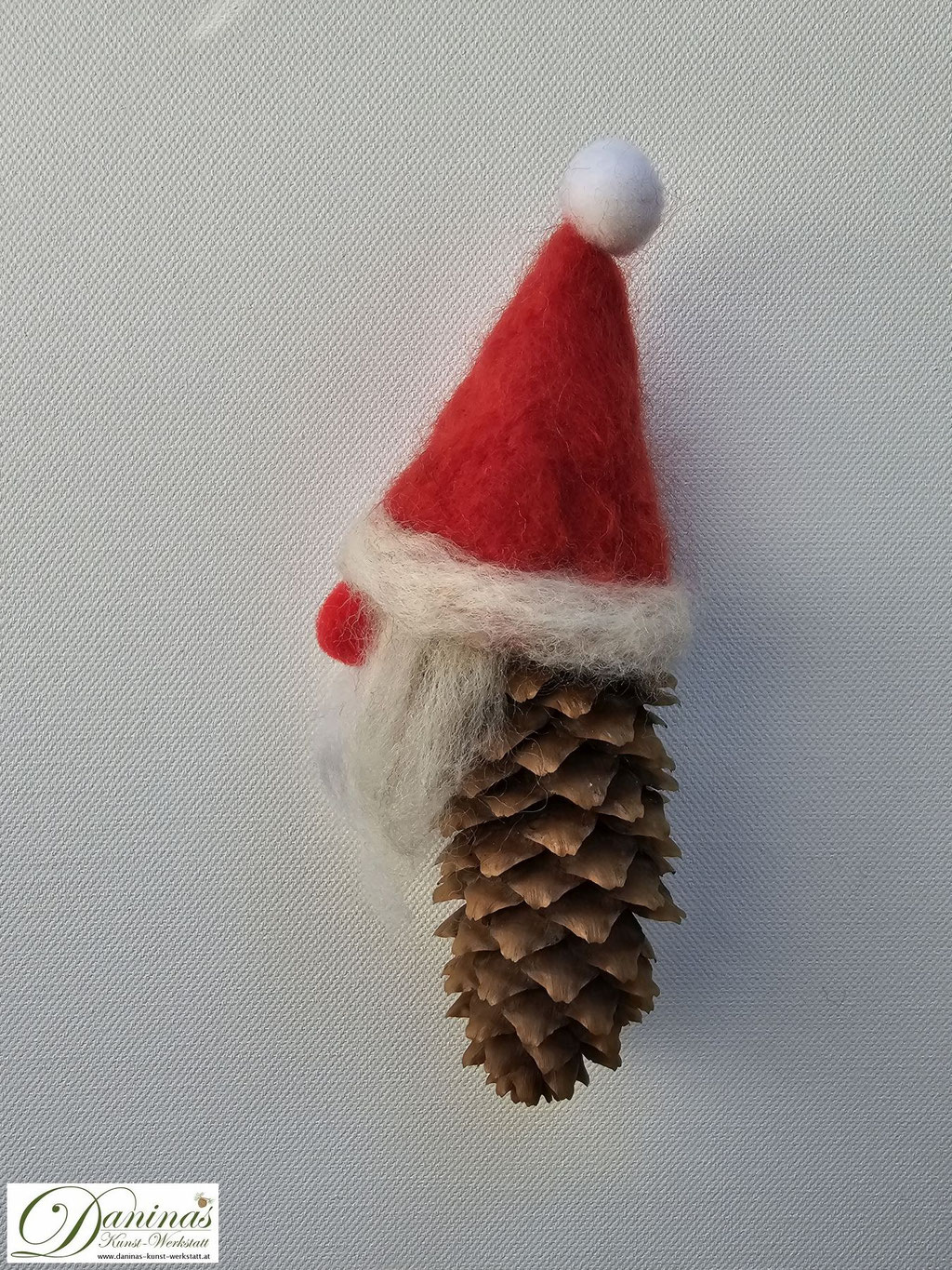 Weihnachtszwerg Rednose, Seitenansicht. Handgefertigte Zwergenfigur aus Fichtenzapfen, mit weißen Haaren und langem Bart aus Wolle, einer rot-weißen nadelgefilzten Wollmütze und einer roten Knopfnase