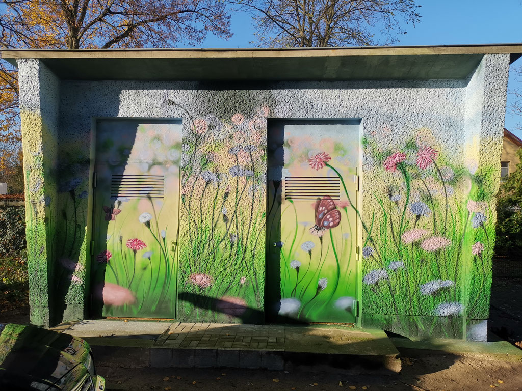Graffiti-Mural auf einem Stromhäuschen zeigt eine farbenfrohe Landschaft mit integrierten Windrädern und Solarpaneelen als Symbol für die Energiewende.