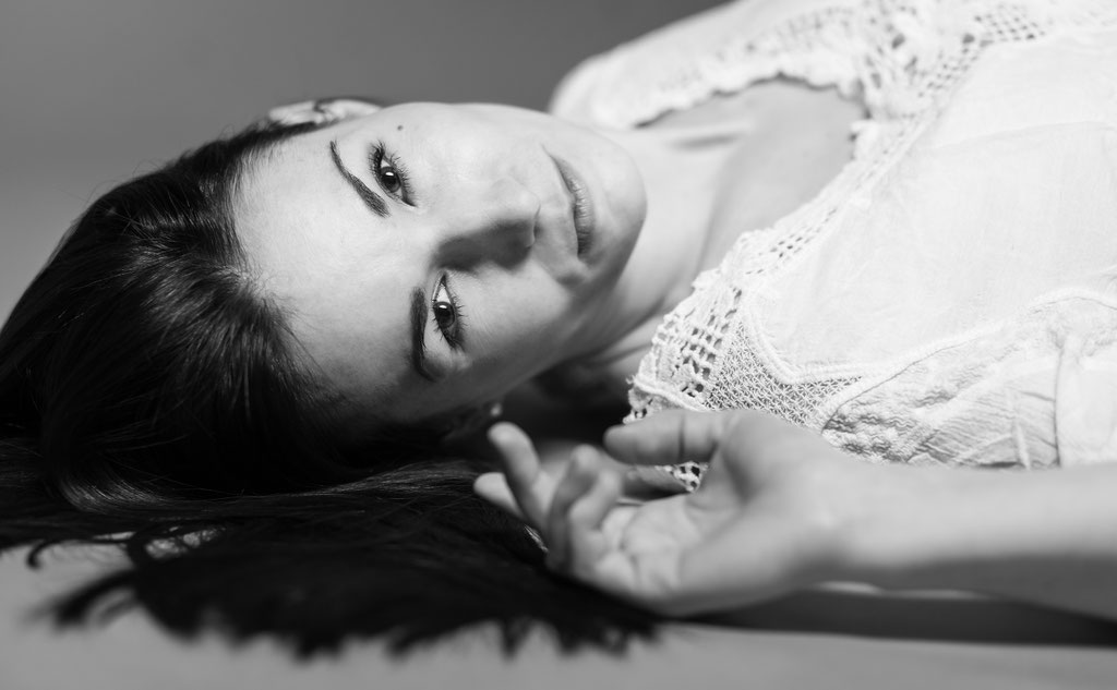 Sinnliches Schwarz-Weiß-Porträt einer Frau, die in verführerischer Gelassenheit auf dem Rücken liegt. Ihr Blick ist intensiv, ihre Hand leicht geöffnet, während das weiche Licht ihre Gesichtszüge und die feinen Details ihrer Spitzenbluse betont.