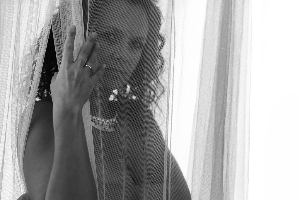 Erotisches Schwarz-Weiß-Porträt einer nackten Frau, die sich hinter einem zarten, transparenten Stoff verbirgt. Ihr intensiver Blick durch den Schleier weckt Neugier – ein Spiel aus Geheimnis und Verführung.