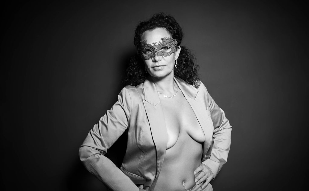 Erotisches Schwarz-Weiß-Porträt einer Frau mit venezianischer Maske, die selbstbewusst in die Kamera blickt. Ihr offener Blazer enthüllt ihre nackte Brust und verleiht dem Bild eine Mischung aus Eleganz, Sinnlichkeit und geheimnisvoller Verführung.