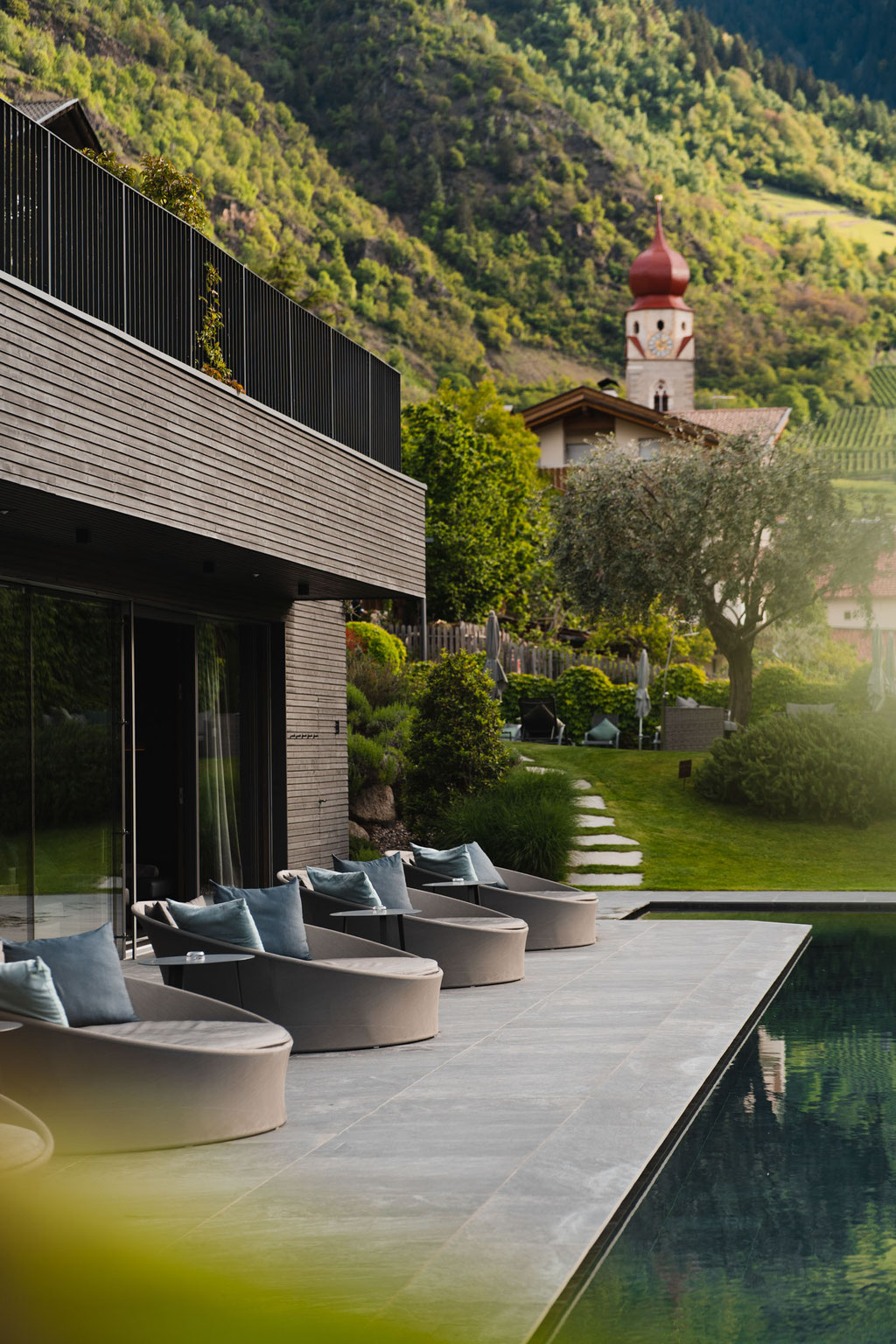 Außenbereich des Hotel Mair am Ort Living in Partschins bei Meran in Südtirol mit stilvollen Lounge-Sitzgelegenheiten am Pool, gepflegtem Garten und Blick auf die historische Kirche sowie die umliegende Südtiroler Berglandschaft.
