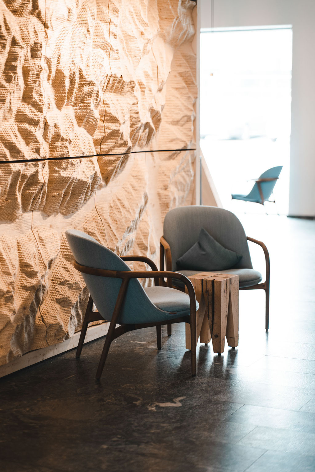Zwei elegante Lounge-Sessel mit kleinem Holztisch stehen vor einer markanten, reliefartig bearbeiteten Holzwand im Garberhof. Die Kombination aus natürlichen Materialien, warmem Licht und klarer Architektur schafft eine ruhige, hochwertige Atmosphäre.