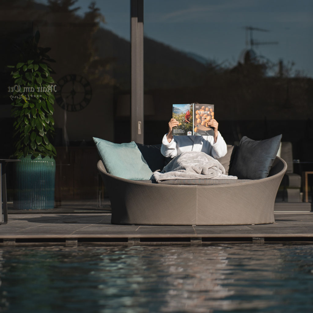 Gast entspannt mit einer Zeitschrift auf einer gemütlichen Loungeinsel am Pool des Hotel Mair am Ort Living in Partschins bei Meran in Südtirol und genießt die ruhige Atmosphäre im Außenbereich.
