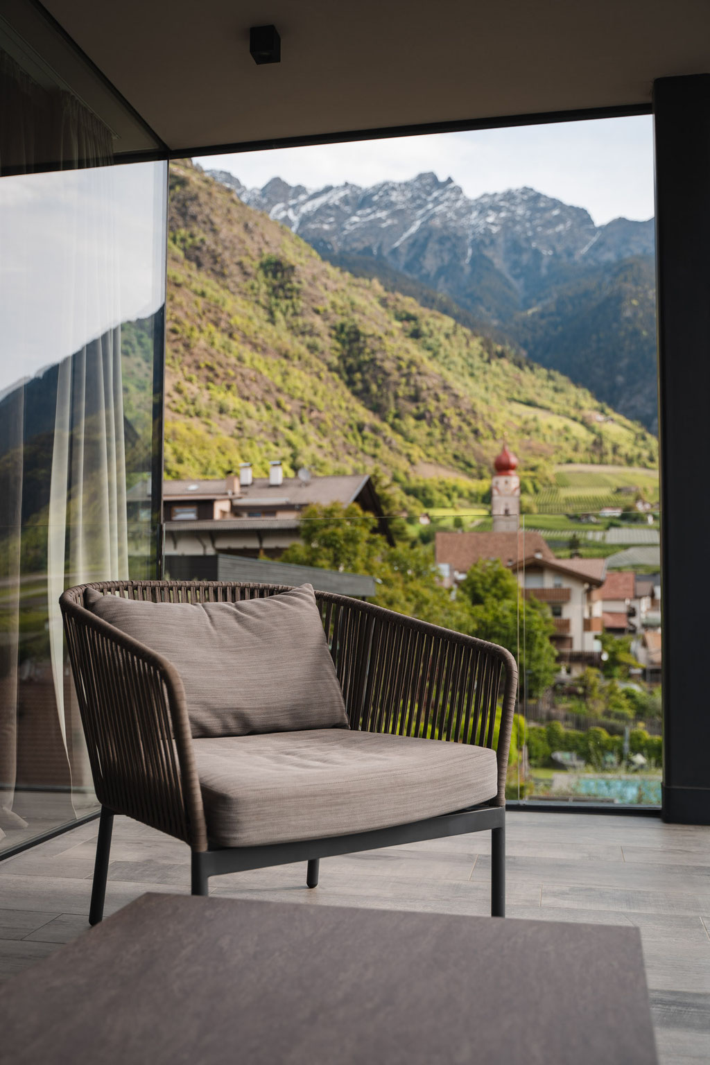 A cosy seating area with a panoramic window at the Hotel Mair in the village of Living, near Partschins, Merano, in South Tyrol. The view sweeps across the village, the striking church tower and the impressive mountain landscape of the Texel Group.