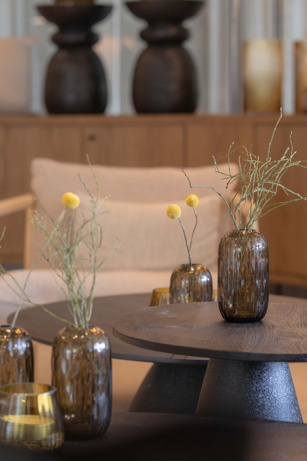 Stilvoll dekorierter Loungebereich mit dunklen Holztischen, Glasvasen mit minimalistischen Zweigen und weichen Sesseln in warmen Naturtönen.