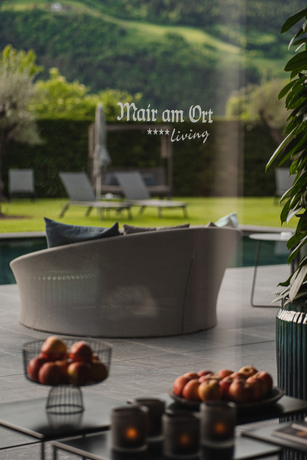A view from the stylish interior of the Hotel Mair am Ort Living in Partschins near Merano in South Tyrol, looking out onto the outdoor pool and the well-tended garden. The hotel logo on the glass façade combines modern design with the hotel’s peaceful, r