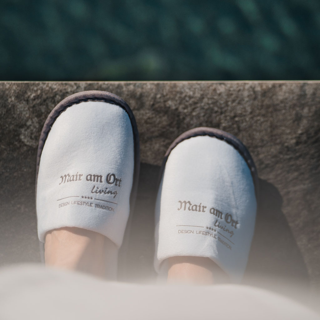 Weiße Hausschuhe mit dem Logo des Hotel Mair am Ort Living in Partschins bei Meran in Südtirol am Beckenrand – ein Detail, das den entspannten Wellness- und Wohlfühlcharakter des Hauses unterstreicht.