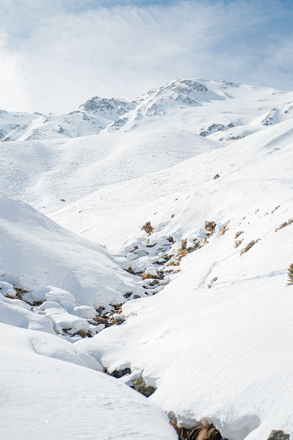 Weite verschneite Berglandschaft mit kleinem Bachlauf im Schnee.