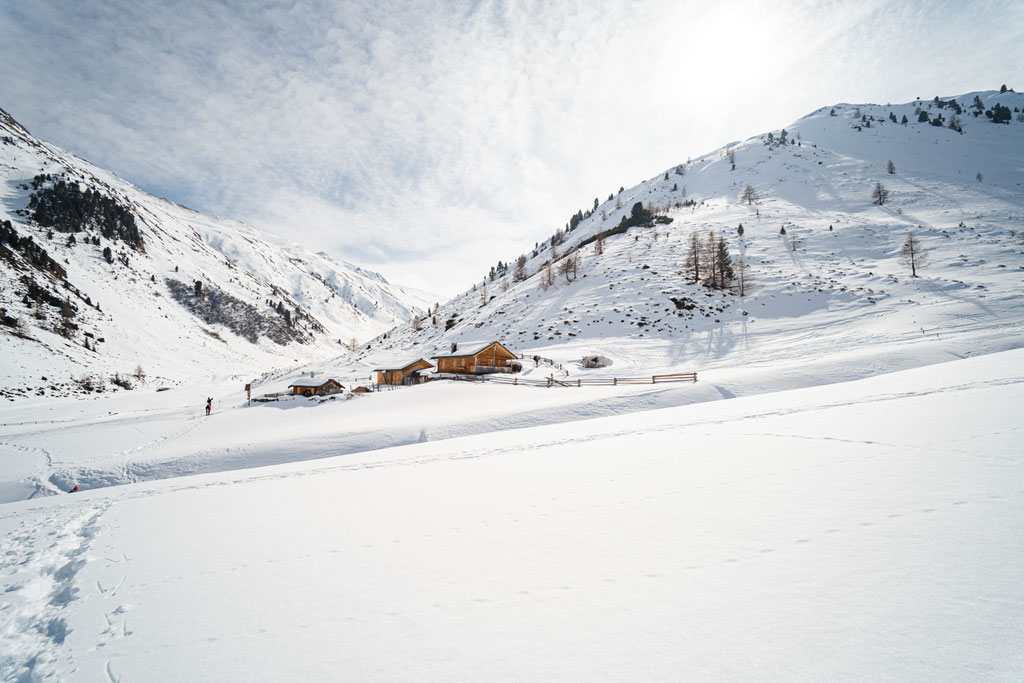 Verschneites Alpental mit Berghütten unter hellem Winterhimmel.