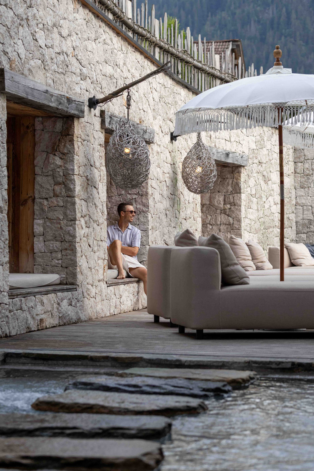 Gemütlicher Loungebereich im Außenbereich des Pure Organic Spa von DER ENGEL im Tannheimertal. Naturstein, Wasser, elegante Daybeds und geflochtene Leuchten schaffen einen stilvollen Rückzugsort mit Blick auf die Bergwelt.