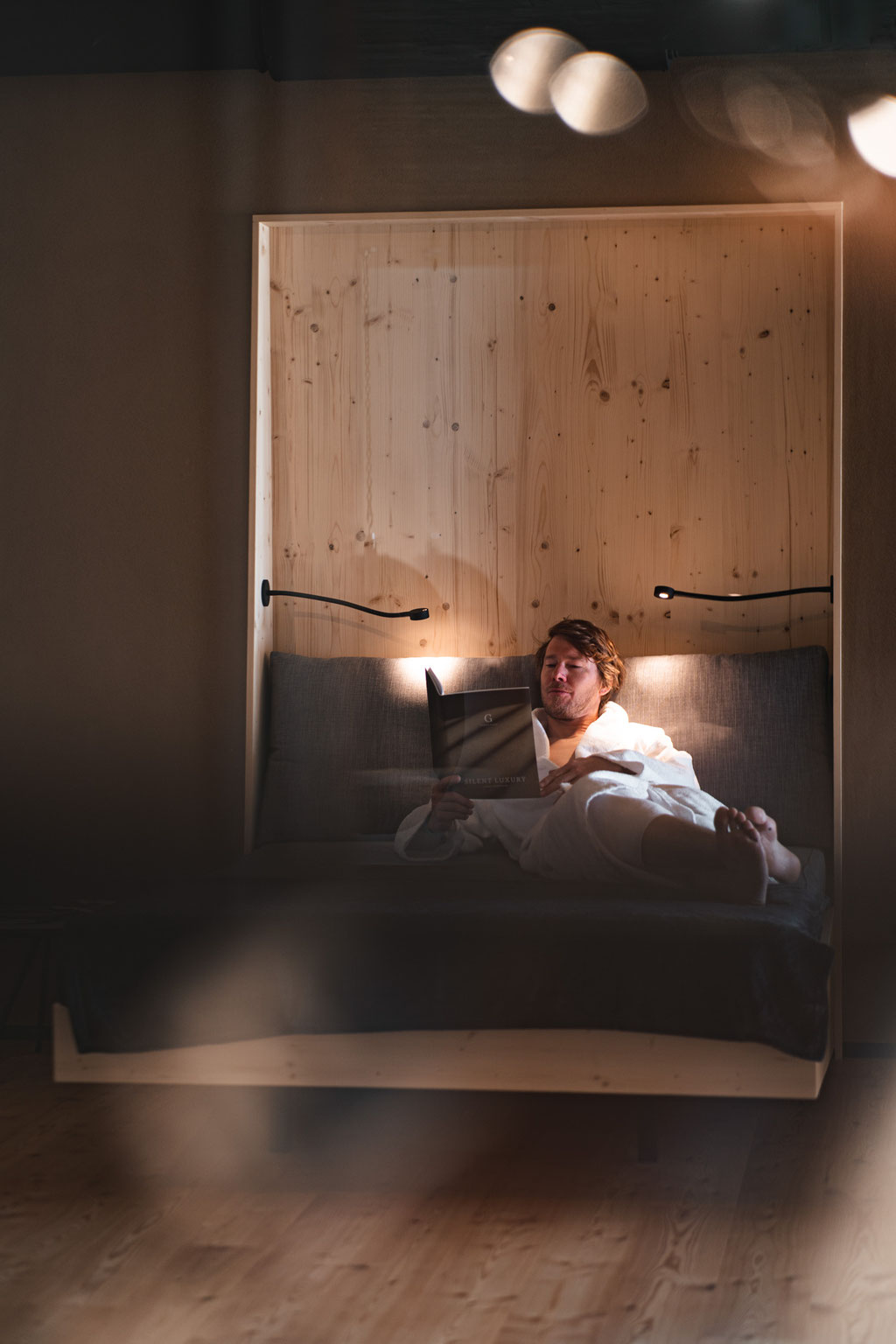 A relaxed hotel guest in a bathrobe is reading in a cosily lit bed.