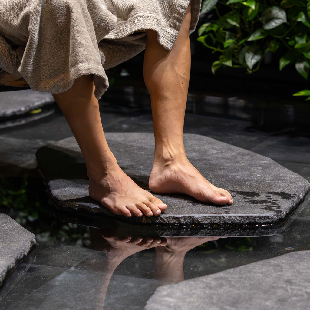 Eine Person mit einem hellen Handtuch um den Körper steht barfuß auf großen Trittsteinen in einem flachen Wasserbecken. Das Wasser spiegelt die Füße wider, während üppige grüne Pflanzen im Hintergrund eine ruhige Spa-Atmosphäre schaffen.