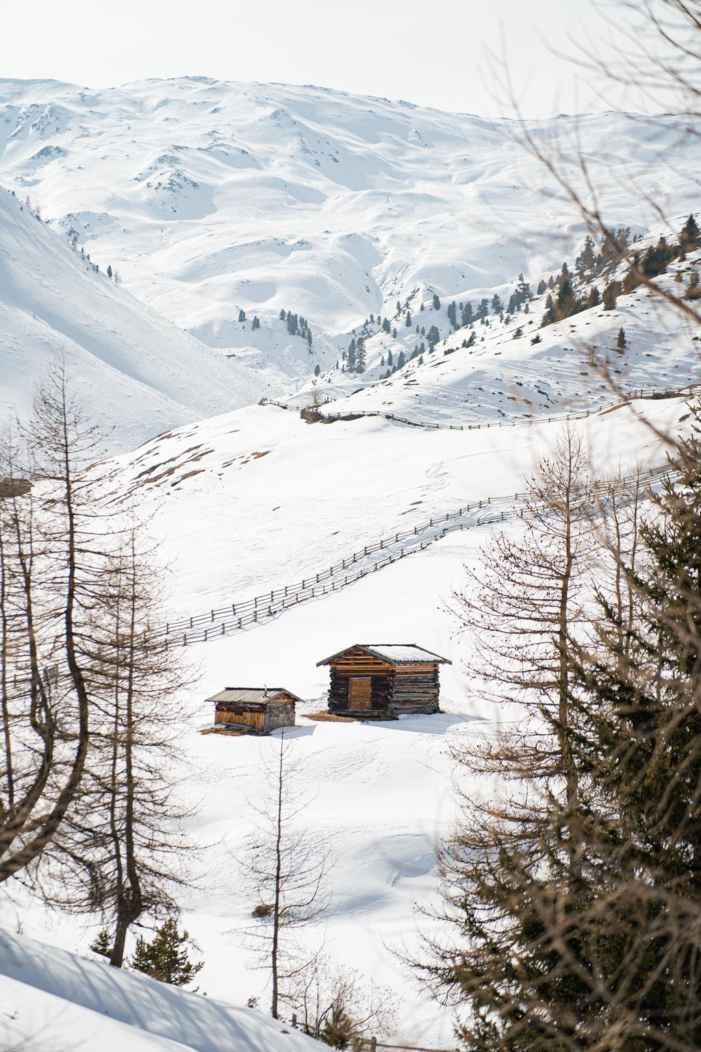 Zwei Holzhütten in verschneiter Berglandschaft mit sanften Alpenhängen.