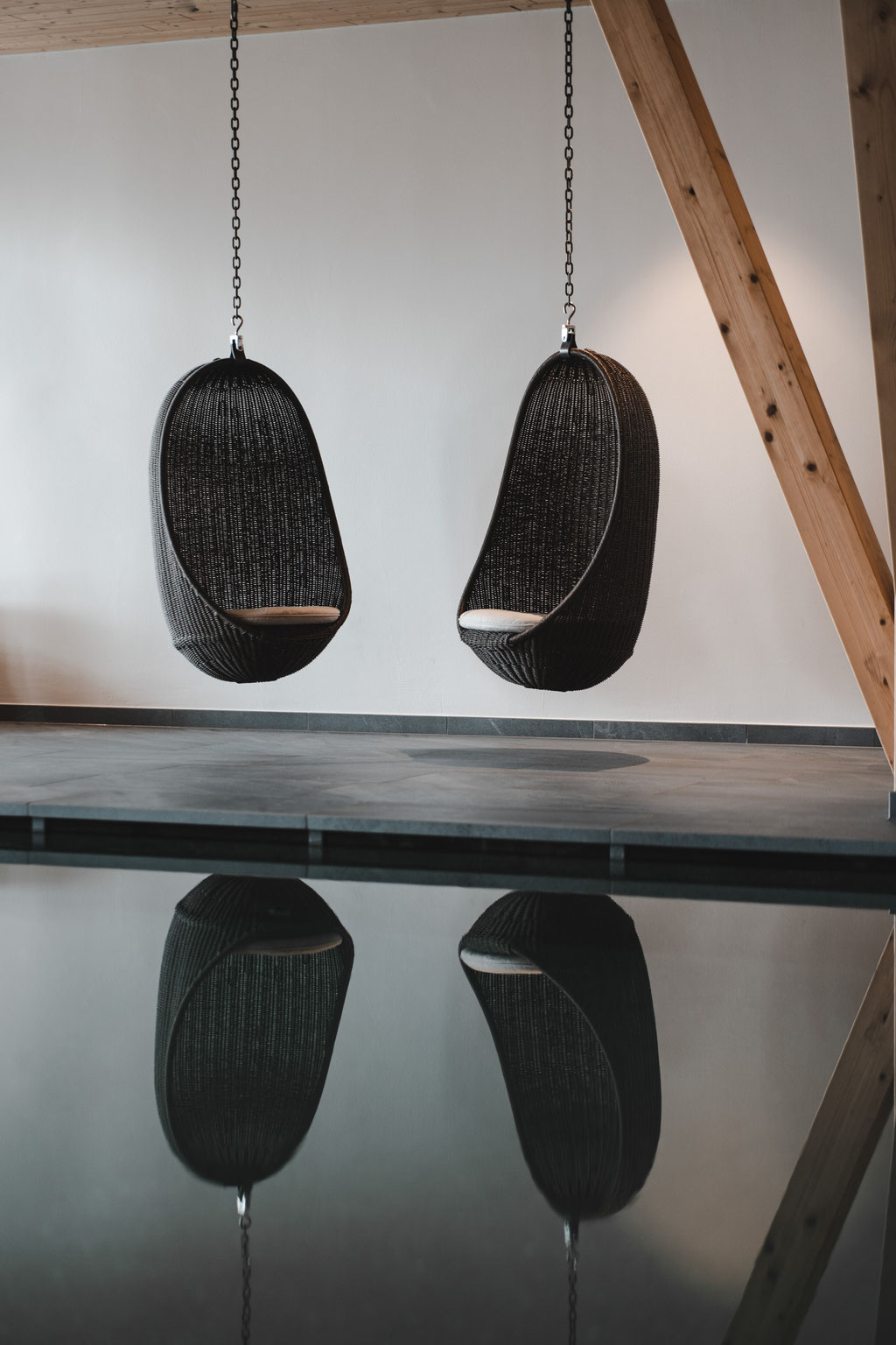 Two hanging wicker armchairs are reflected in the calm water of an indoor pool.