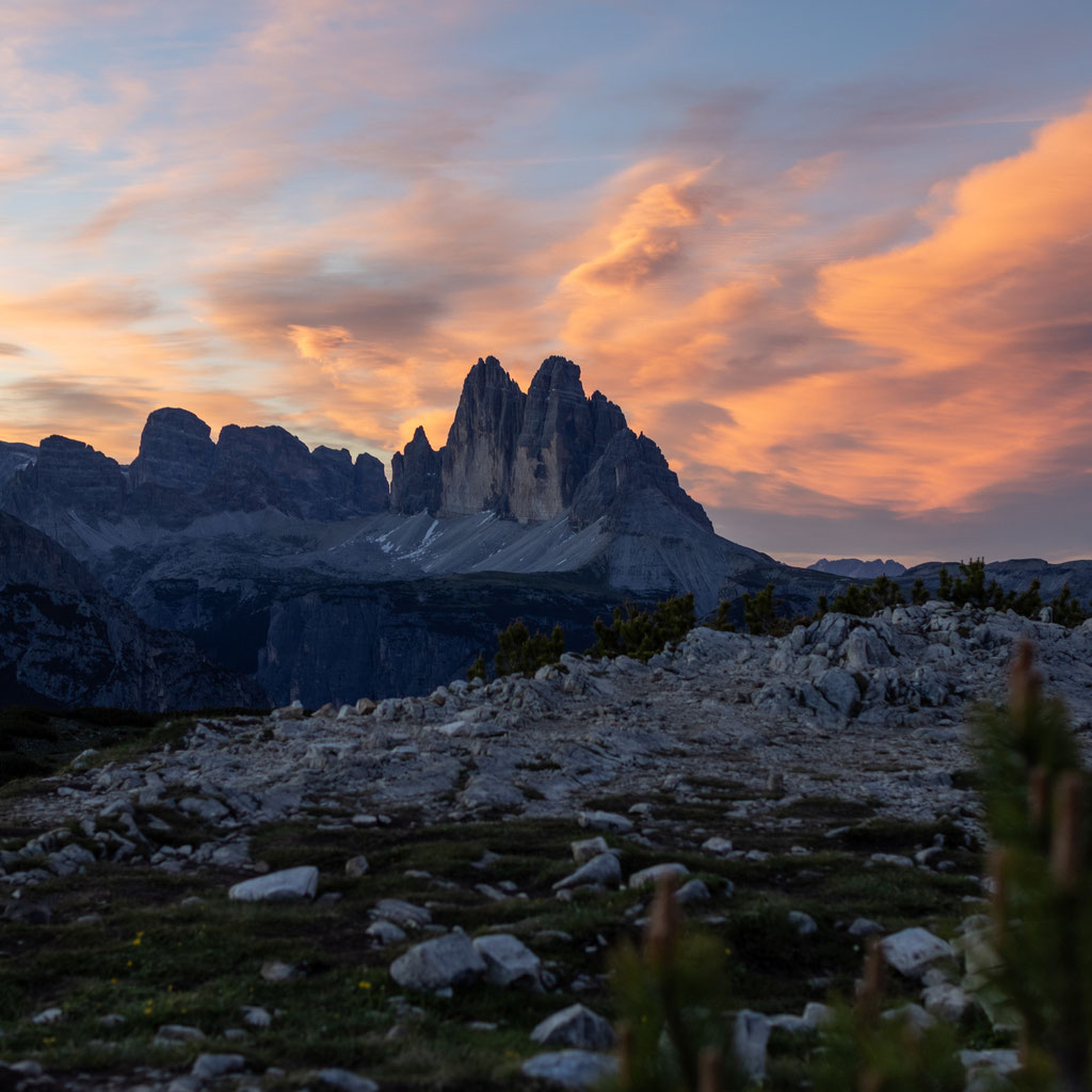 Die Drei Zinnen in den Dolomiten erheben sich vor einem spektakulären Himmel, der in kräftigen Orange-, Rosa- und Blautönen leuchtet. Im Vordergrund erstreckt sich eine felsige Almlandschaft, die die ruhige und beeindruckende Abendstimmung unterstreicht.